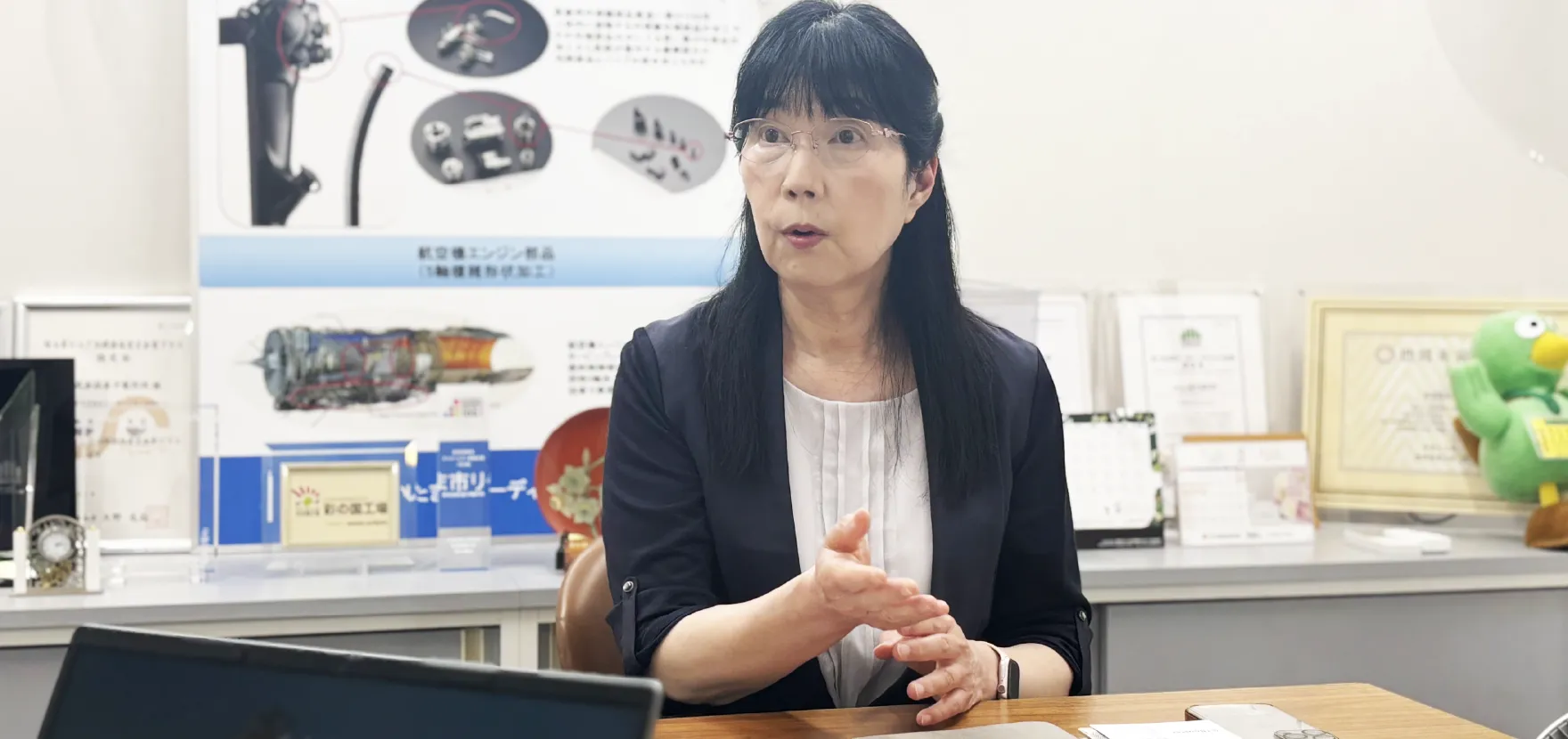 オフィスで机に向かい、身振り手振りを交えながら話している女性。背景には航空機エンジンのパネルが見える。